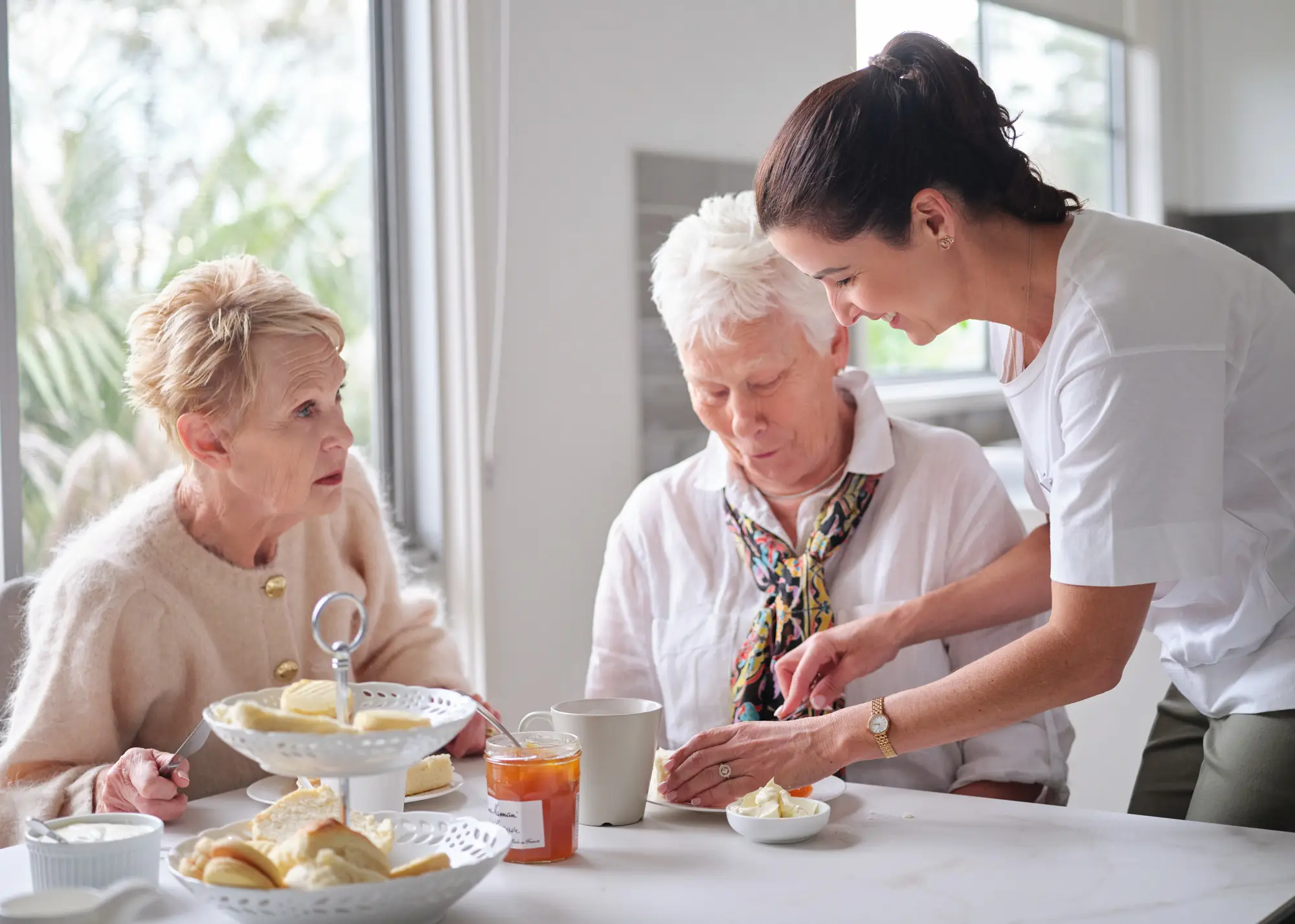 A carer helping two older women with morning tea at the kitchen bench.